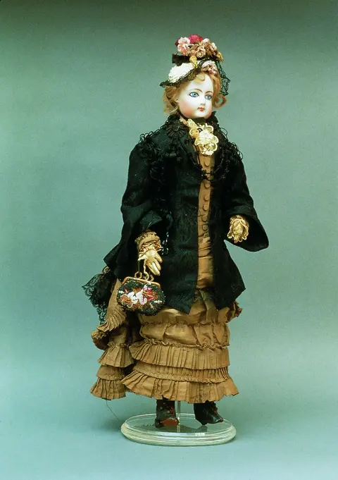 A full-length bisque porcelain doll dressed in an elaborate 1870s dress with a flowered hat
